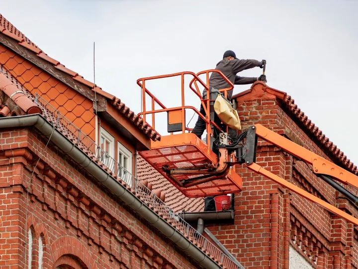 Roofer on a cherry picker repairing a tiled roof on a red brick building