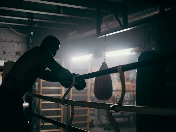 Moody silhouette of a boxer leaning in a dimly lit gym with punching bag and boxing ring.