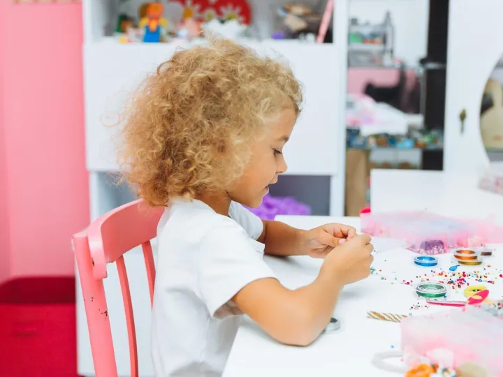 Cute little blonde girl making bead jewelry at a table in th