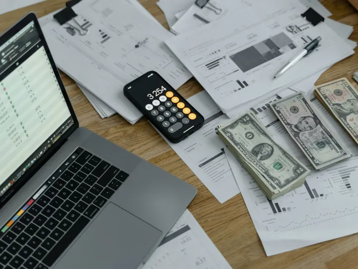A desk with financial documents, currency, a laptop, and phone calculator.