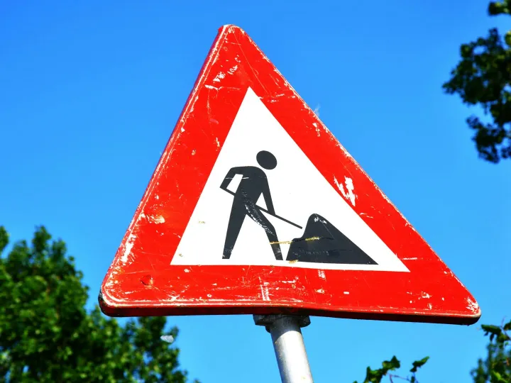 A red and white roadwork warning sign with a silhouette figure, set against a clear blue sky.