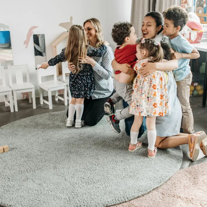 Young children hugging two nursery practitioners in a playroom