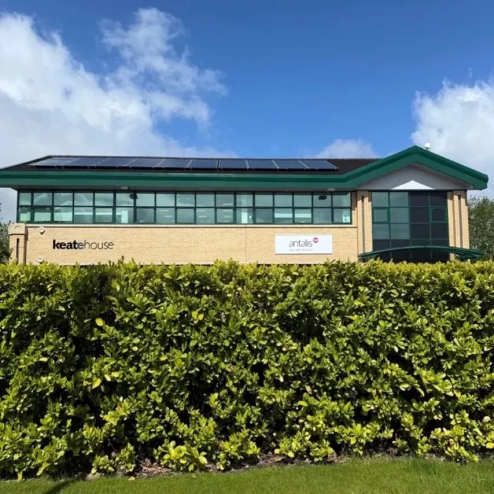 Brick office building with green window frames and rooftop solar panels behind a tall hedge