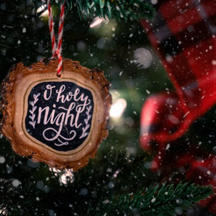 Wooden bauble with O Holy Night inscription hanging on green Christmas tree decorated with red ribbon on snowy day