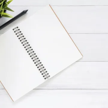 Open spiral notebook on white desk with black pen and green plant. Ideal for planning.