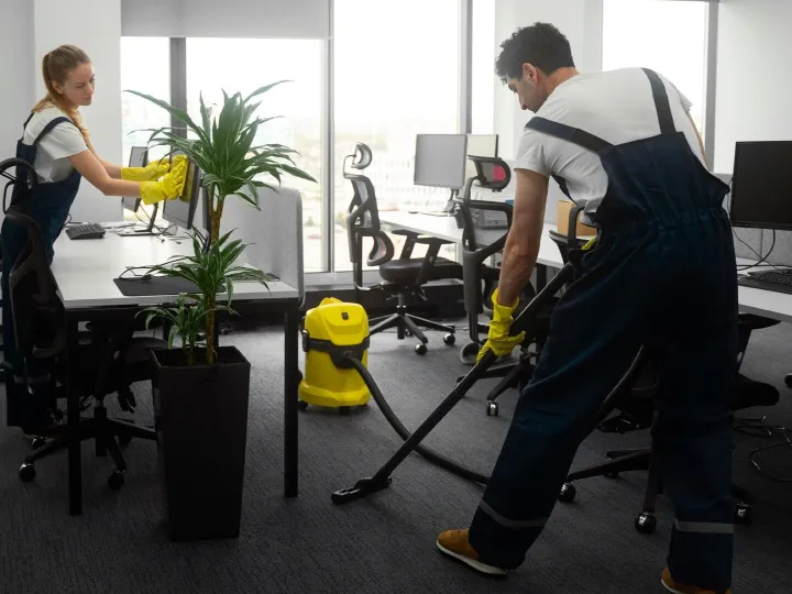 Cleaner vacuuming an office carpet beside a bright window