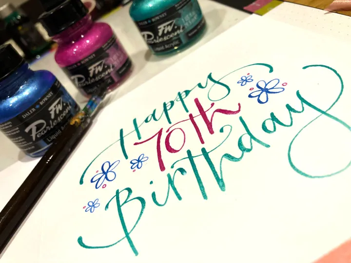 Happy 70th birthday card lettered in blue and pink beside pots of ink