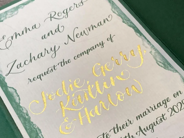 Hand lettered wedding invitation with gold foil calligraphy names and a green watercolour border