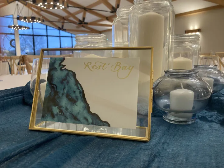 Hand lettered gold wedding place card with a watercolour coastal map, framed among candles