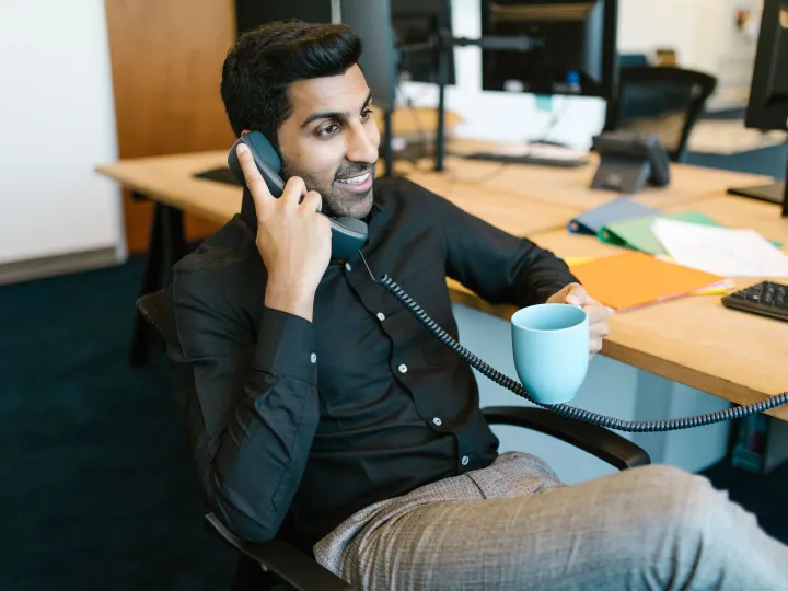 A man smiling as he answers a desk telephone, holding a mug