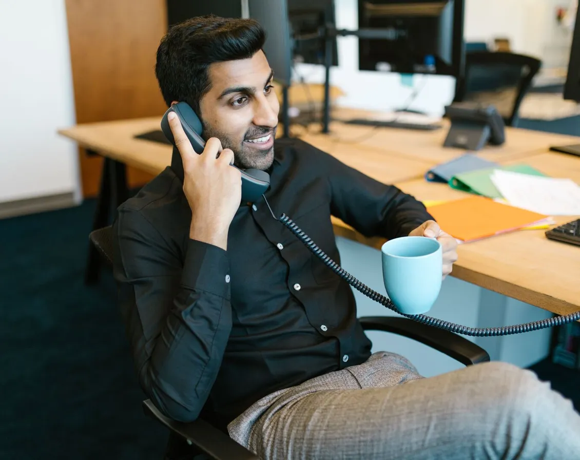 A man smiling as he answers a desk telephone, holding a mug