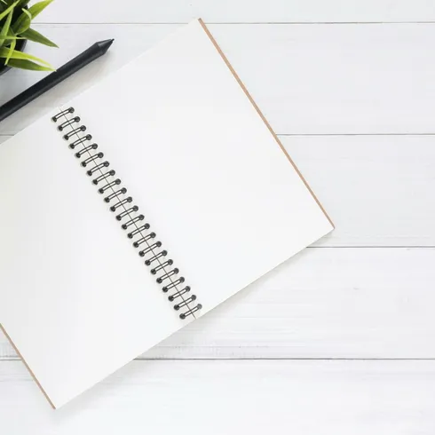 Open spiral notebook on white desk with black pen and green plant. Ideal for planning.
