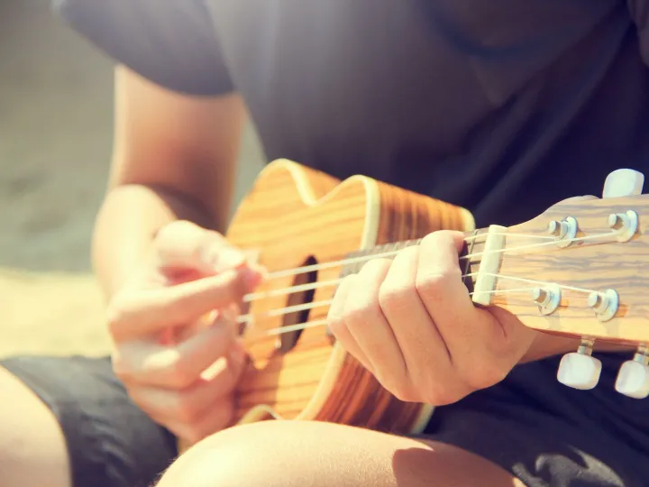 Close-up of a man's hands strumming a ukulele outdoors on a sunny day.